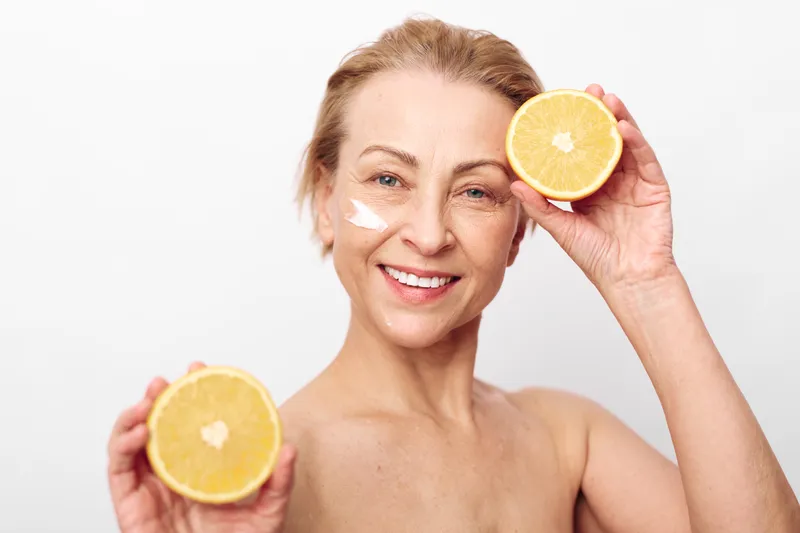 Smiling mature woman with natural skin holds two halves of a fresh citrus fruit near her face. Visible cream on cheek symbolizes skincare, wellness, and natural beauty concept on white background.