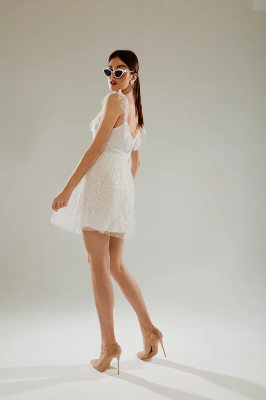 Young woman in a white lace dress and sunglasses poses confidently in high heels on a neutral background. Concept of woman beauty fashion and modern style.