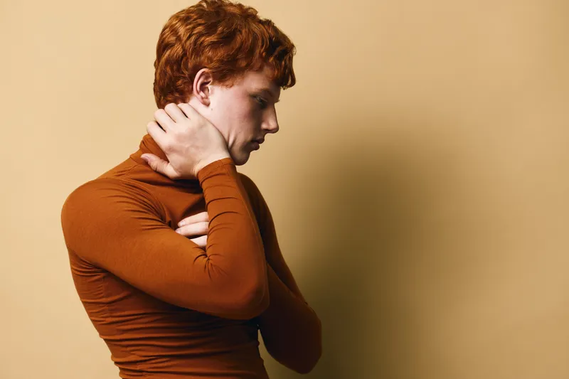 Redhead man in orange turtleneck posing with hand on neck looking down thoughtful isolated on beige background. Side profile of young male model with calm expression and casual style.