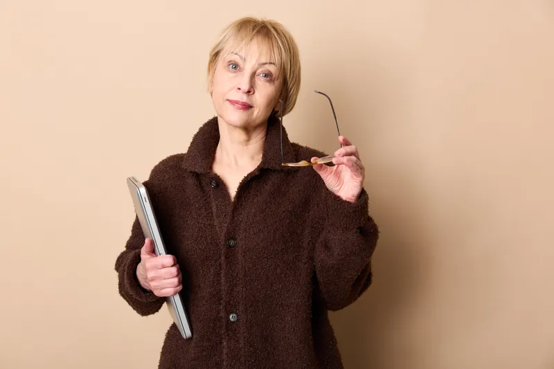 Elderly woman indoors, studio portrait, holding tablet and glasses, wearing a dark coat, facing camera with calm thoughtful expression, soft light on beige background