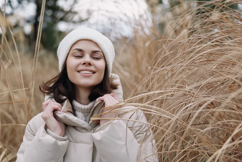 Woman in winter jacket and beanie smiles among tall dry grasses in an outdoor field, soft natural light creates a cozy, calm portrait suitable for seasonal lifestyle and nature themes.