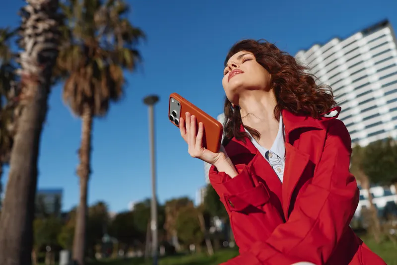 A smiling woman in a red jacket enjoys bright sunshine outdoors, holding a smartphone up in a park with palm trees and a clear blue sky.