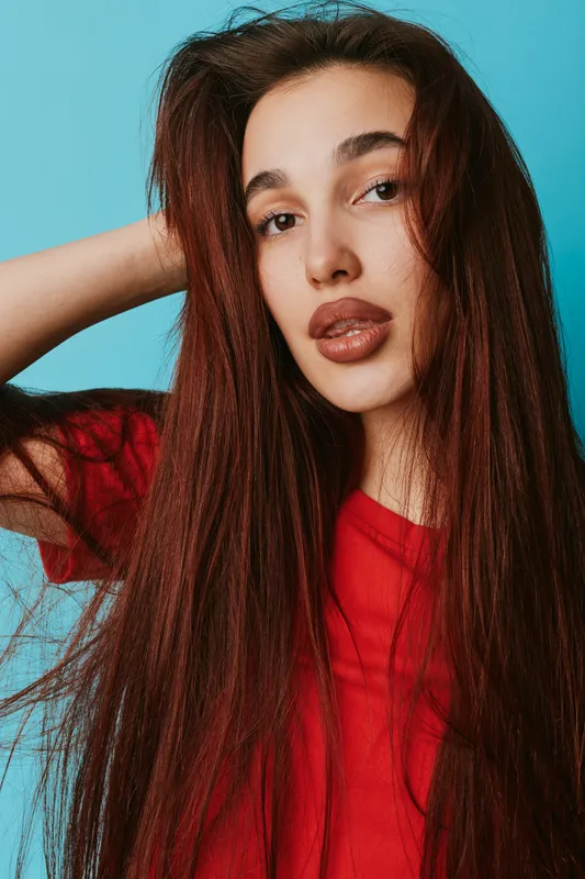 Young woman with long brown hair wearing red shirt posing with hand behind head against blue background. Portrait of female model with natural makeup and confident expression.
