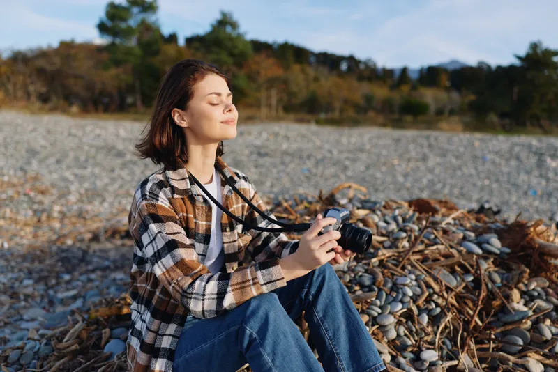 Female photographer sitting on a rocky shore with a camera, basking in sun and breeze. Casual plaid shirt, denim jeans, natural light, tranquil seaside moment, outdoor creativity, focus during a