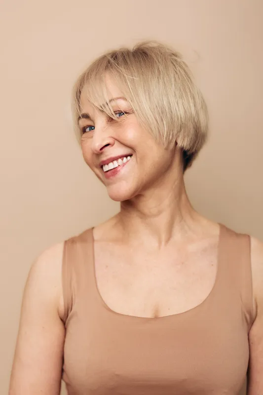 Smiling middle aged woman with short blonde hair wearing sleeveless beige top poses against neutral background. Concept of happiness, confidence, natural beauty, and mature lifestyle.