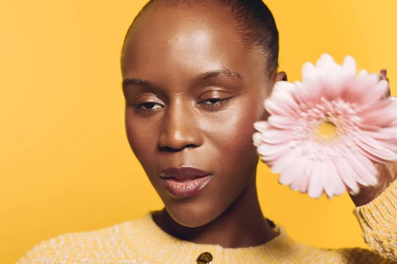 Close-up of a dark skinned African woman holding a pink flower near her face against a yellow background. Concept of beauty, skincare, nature, and natural glow.