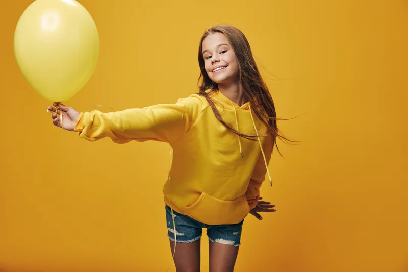 Girl smiling and holding yellow balloon in hand, wearing yellow hoodie and denim shorts standing against yellow background. Child posing with joy and casual look in studio.