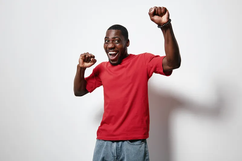 African American man with dark skin wearing red t shirt and jeans smiling and celebrating with raised fists on white background. Happy male person expressing joy and excitement.