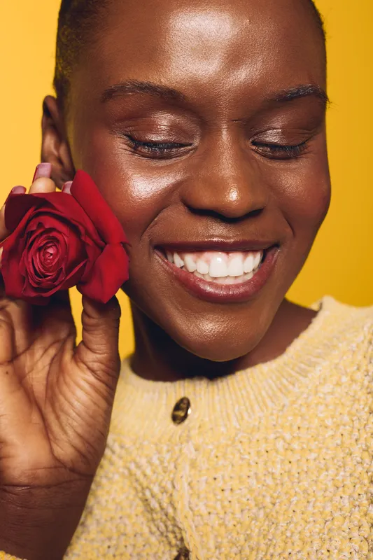Close-up of smiling black woman with dark skin holding red rose near face against yellow background. Concept of happiness, beauty, and natural skincare for African ethnicity.