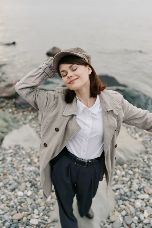 Young woman in casual beige jacket and cap enjoys fresh air with closed eyes near rocky shore. Concept of relaxation, nature, freedom, and peaceful outdoor lifestyle by the sea.