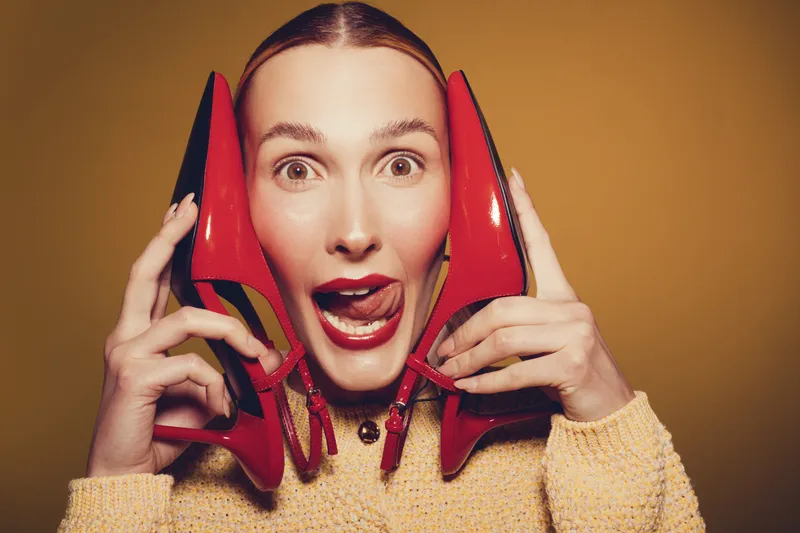 Young woman with red lipstick holds red high heel shoes near face, making playful expression. Yellow background and warm sweater create vibrant, fun fashion concept.
