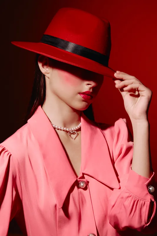 Fashion woman in pink blouse and red hat hides face with brim. Pearl necklace and blush makeup highlight creative beauty look. Editorial style with neon red background.