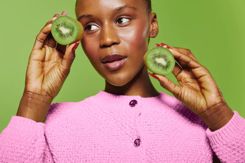 Young black woman with dark skin holds two halves of kiwi fruit near face. She wears a pink sweater and poses against a green background. Concept for healthy lifestyle and natural beauty.