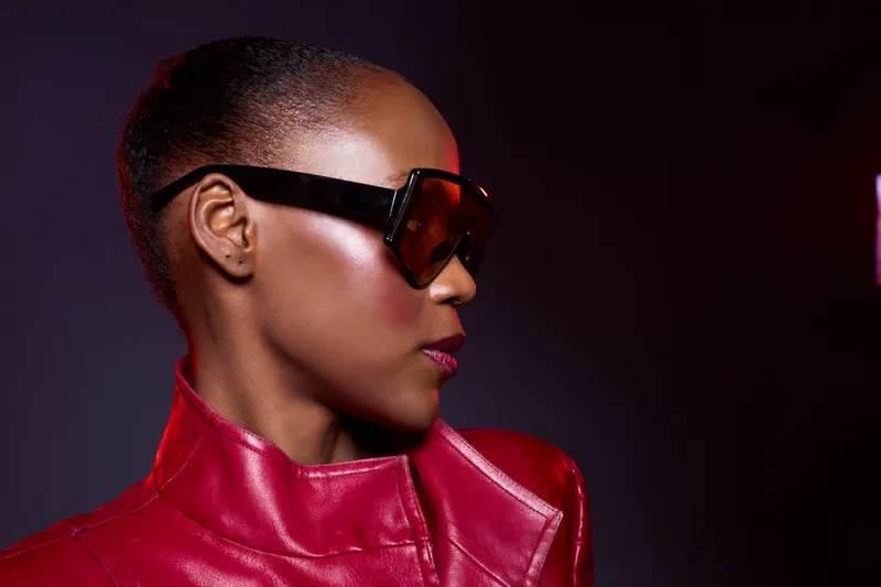 Stylish black woman with dark skin wears large sunglasses and a red leather jacket. She poses confidently against a dark background, showcasing fashion and modern elegance.