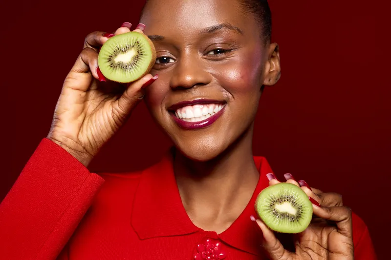 Smiling black woman holds fresh kiwi fruit halves near face against red background. Concept of healthy eating, natural skincare, and joyful lifestyle with dark skin female model in vibrant clothing.