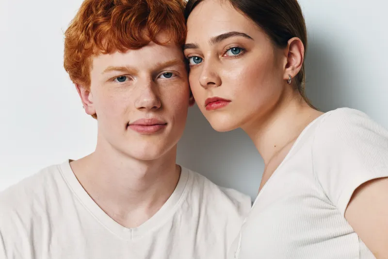 Young couple with serious expressions posing together in casual white shirts against a neutral background. Closeup portrait showing natural skin and eye contact.