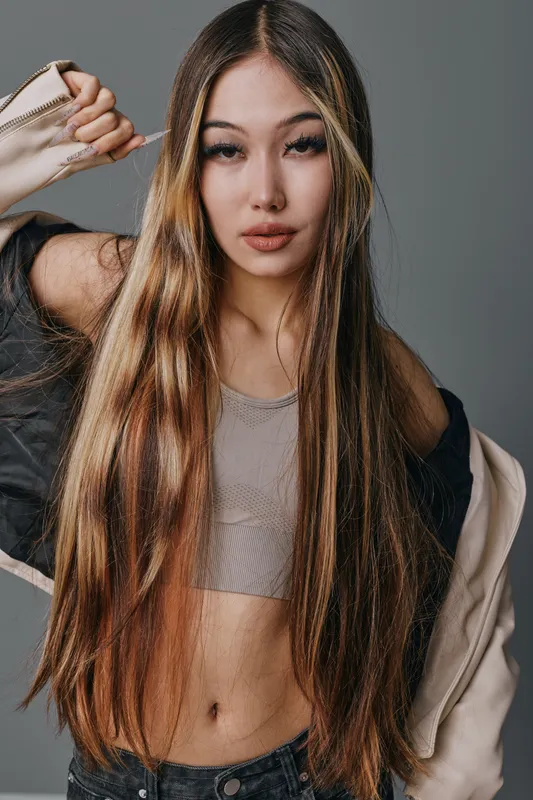 Young woman with long hair posing indoors wearing casual top and jacket with confident expression and natural makeup on neutral background for lifestyle photo.