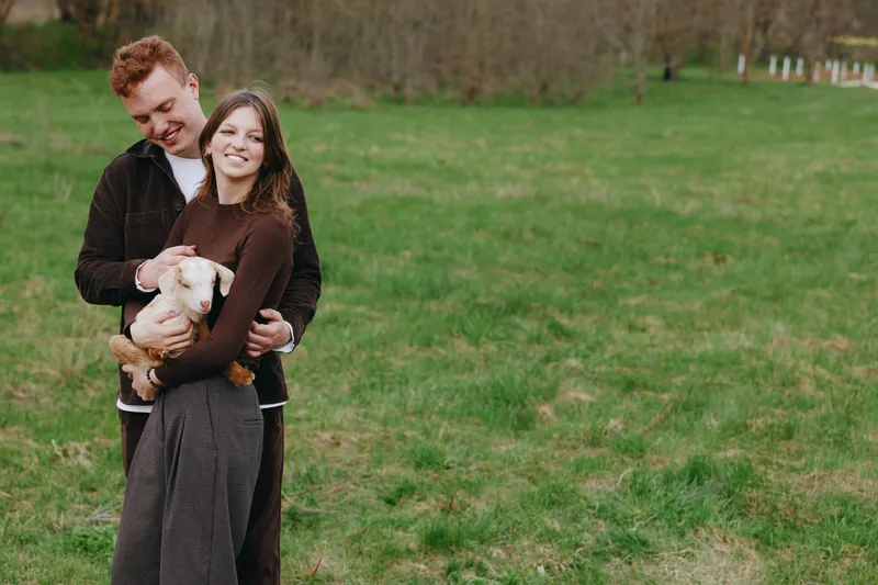 Joyful couple outdoors with a goat kid, embracing in a grassy field, warm sun, relaxed pose, candid moment, natural colors, rural backdrop, simple happiness, wholesome lifestyle