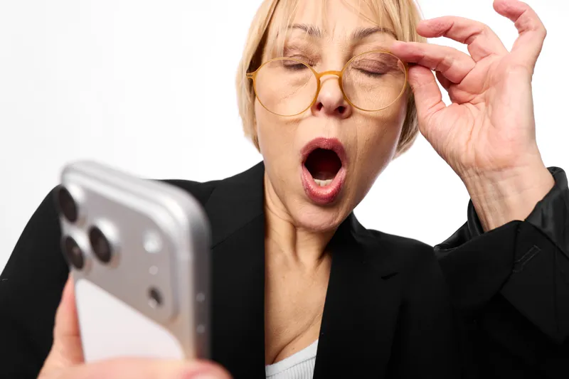 Elderly woman with glasses holding smartphone for selfie, surprised open mouth, hand near eye, modern technology, home communication, studio light, portrait