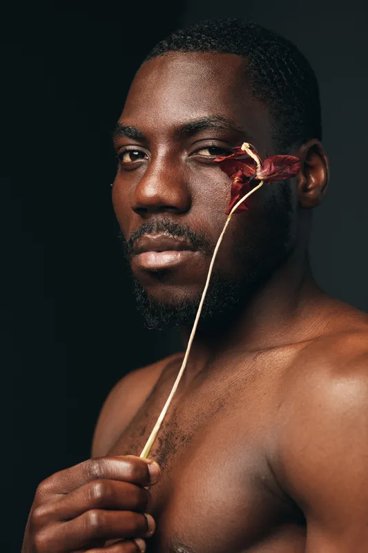 Black man with dark skin holding dried flower near face in creative conceptual visual style portrait. African ethnicity, artistic expression and natural beauty with bare shoulders on dark background.