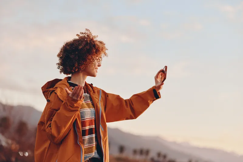 Woman lifestyle outdoor in orange jacket, curly hair, smiling profile, arm raised as if taking a selfie, warm sunset light above mountains, calm autumn travel mood, natural scene