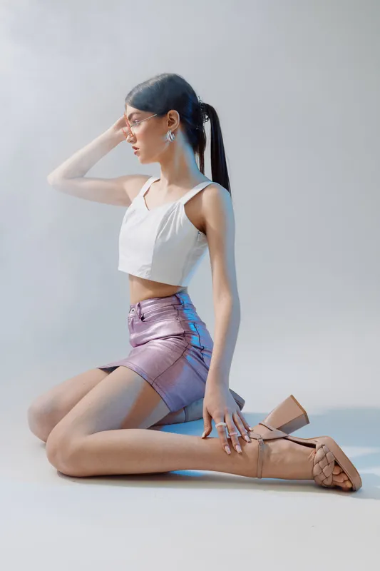 Young woman in a white crop top and shiny purple skirt poses kneeling on the floor. Her hair is tied back and she looks to the side in a studio with soft lighting. Concept of beauty fashion.