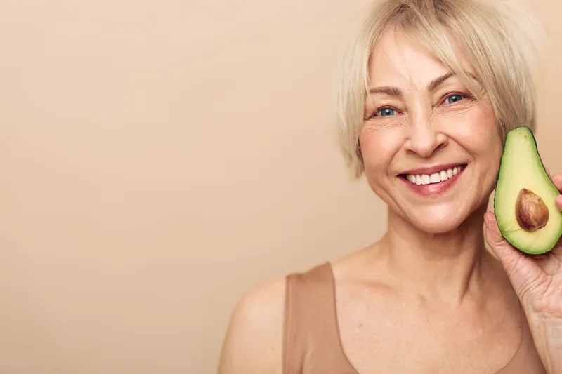 Smiling mature woman with blonde hair holds fresh avocado half near her face on beige background. Concept of healthy lifestyle, nutrition, wellness, and natural beauty for older adults.