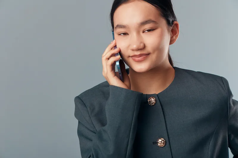 Young woman talking on mobile phone with slight smile wearing gray blazer against plain background. Communication, businesswoman and technology for networking or casual conversation indoors.