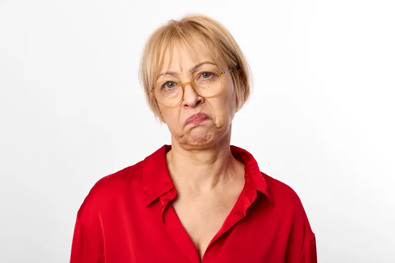 Woman senior with glasses applying white face cream, skincare mask on cheeks, red blouse portrait, white studio background, soft lighting, wellness concept