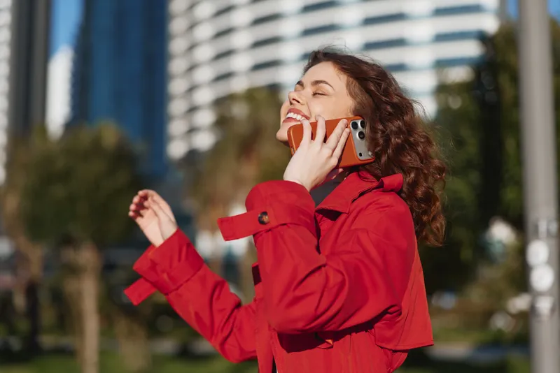 Young woman with curly hair wearing a red coat talks on a smartphone in a bright city park, smiling as modern buildings rise behind her.