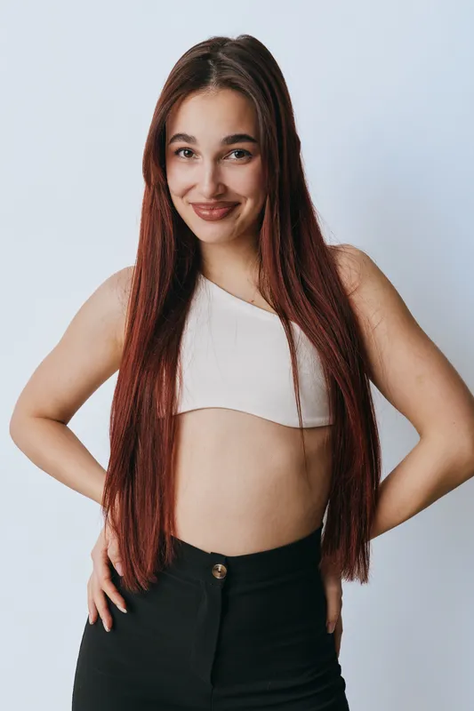 Young woman with long red hair wearing a white crop top and black pants posing with hands on hips against white background. Smiling confident female model standing indoors.