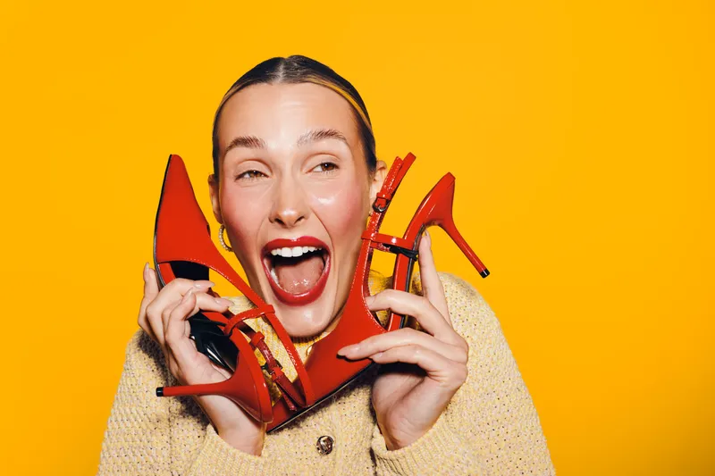 Happy woman with red high heels holds shoes near face smiling widely on yellow background. Concept of fashion, style, footwear, and joyful emotions in studio lighting.
