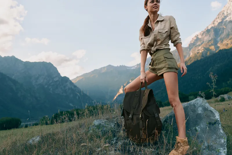 A young woman stands on a rocky meadow with a backpack in a rugged mountain landscape, ready for a hike and outdoor adventure.
