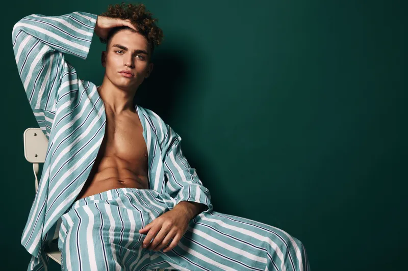 Young man with curly hair wearing striped pajamas sitting on chair against dark green background, posing with hand on head and relaxed expression in studio lighting.