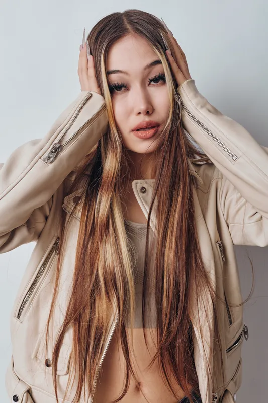 Young woman with long straight hair and braids posing in beige leather jacket and crop top, looking expressive against white background indoors in studio.