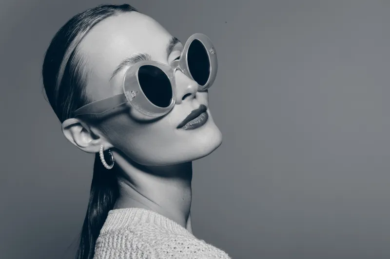 Young white woman with fair skin wearing round sunglasses and hoop earrings looks away, wearing knitted sweater on grey background. Concept of fashion and style.