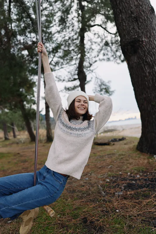Woman on a swing in an outdoor park, wearing a cozy sweater and beanie, smiles as she enjoys a breezy autumn afternoon among trees.