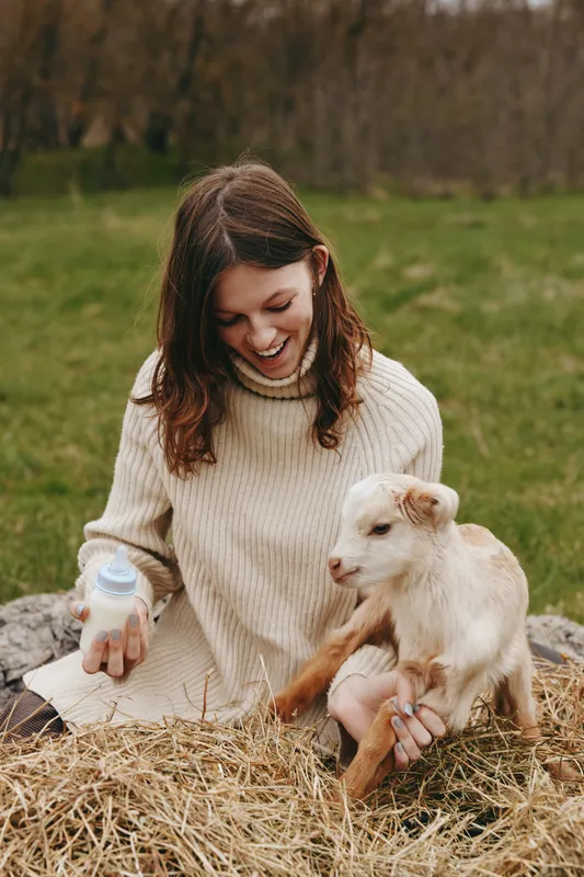 Bright outdoors scene with a smiling woman feeding a goat kid from a bottle, cozy sweater, sitting by a hay bale, warm natural tones, tender lifestyle moment in rustic countryside