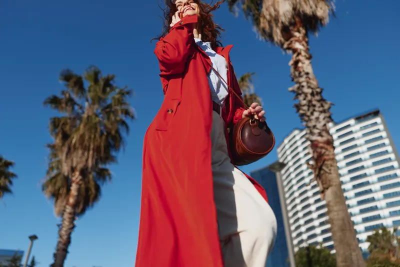 Young woman wearing a long red coat walks confidently outdoors on a sunny day near palm trees and modern buildings, carrying a round brown handbag.
