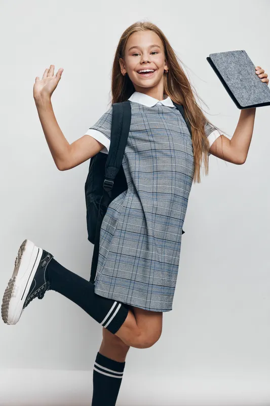 Girl student jumping and smiling with backpack and tablet in hand for education, learning and school concept. Child wearing uniform with happy expression and energetic pose on light background.