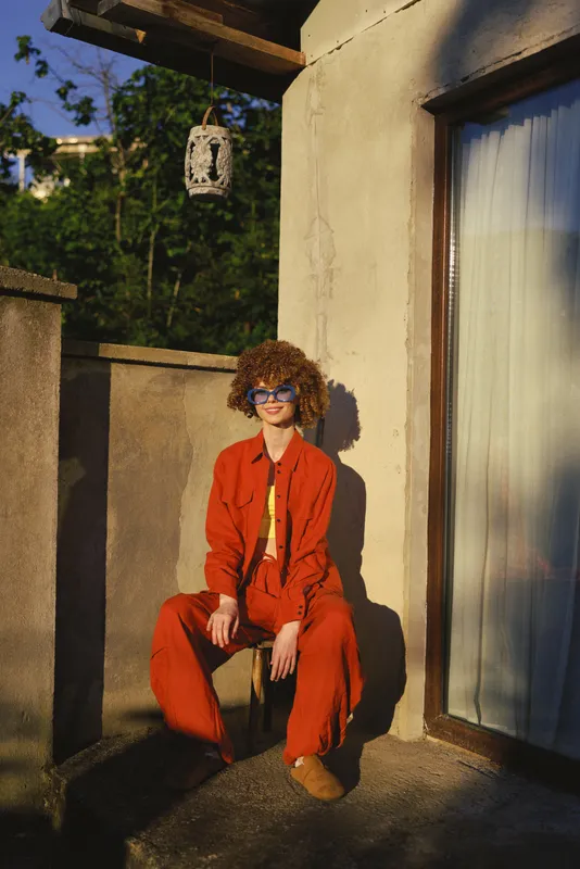 Confident woman with curly hair wears bright red suit and sunglasses, seated on a sunlit terrace, urban lifestyle portrait in warm daylight, relaxed casual pose, stylish energy