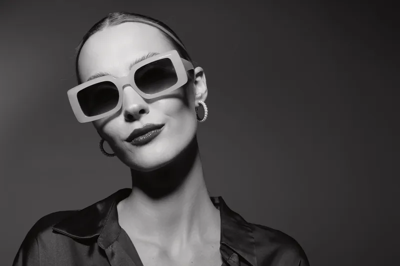 Stylish caucasian woman with pale skin wearing large sunglasses and dark lipstick poses confidently against black background. Concept for fashion and modern elegance.
