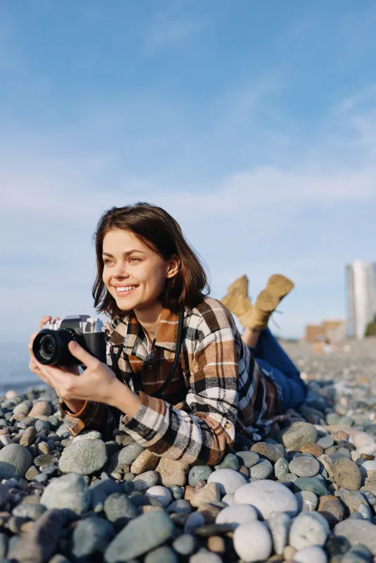 Female photographer lying on pebbles at the beach, smiling while holding a camera for outdoor lifestyle photography and creative seaside moments in bright daylight.