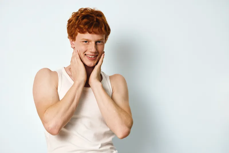 Young man with red hair and fair skin wearing sleeveless white shirt smiling and touching face against plain white background in studio portrait.