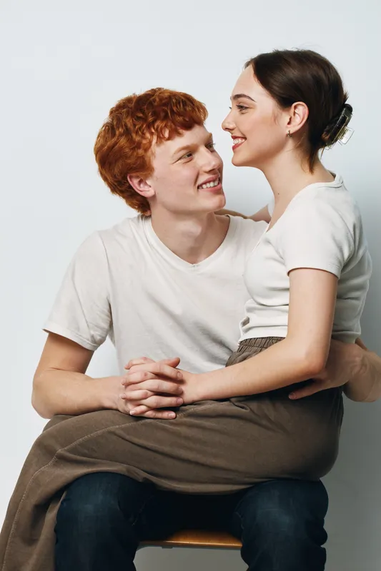 Couple in love with woman sitting on man’s lap smiling and looking at each other for relationship, affection and intimacy in casual clothes in studio.