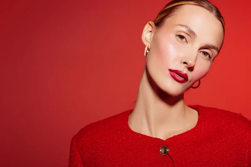 Portrait of a confident woman with glossy skin and red lipstick wearing a red sweater against a red background. Concept of beauty, fashion, and makeup with vibrant colors.
