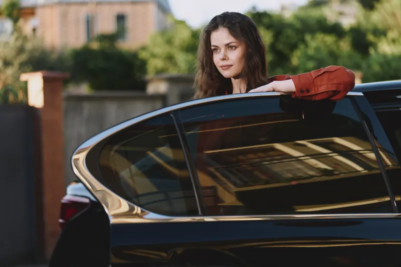 Young woman leans on a sleek car outdoors, casual lifestyle portrait with a thoughtful gaze, illuminated by natural daylight in an urban setting, relaxed mood