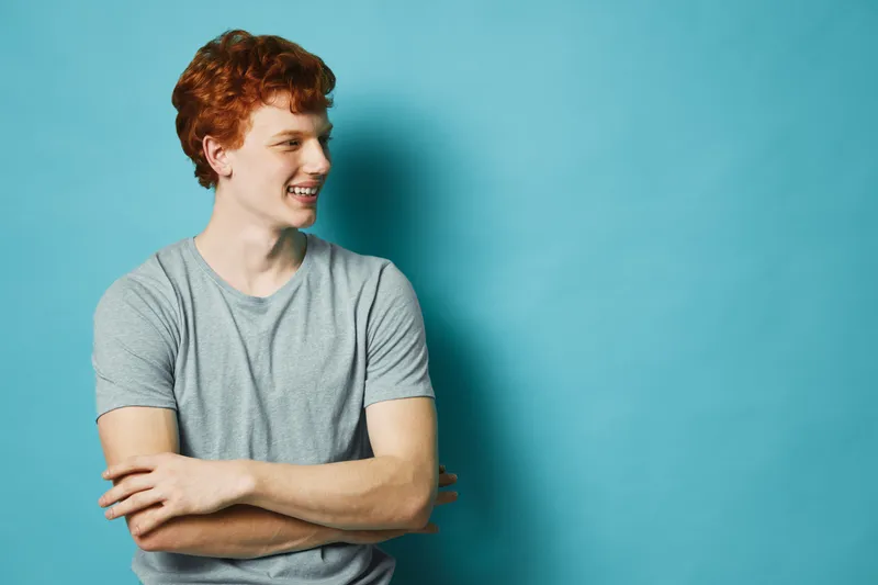 Young man with red hair and freckles wearing gray t shirt standing with crossed arms smiling and looking sideways against blue background. Casual portrait with positive expression.