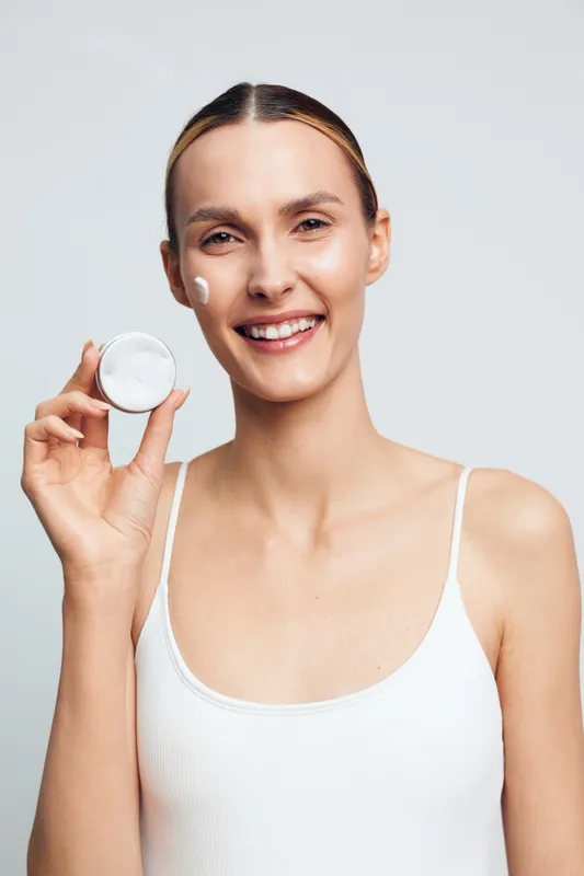 Smiling woman shows skincare routine, holding cotton pad for facial hygiene and beauty care, isolated on light studio background, close up portrait with clean complexion.