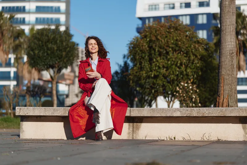 Young woman in red coat sitting on stone bench outdoors smiling holding smartphone with city buildings and trees in background under clear sky.